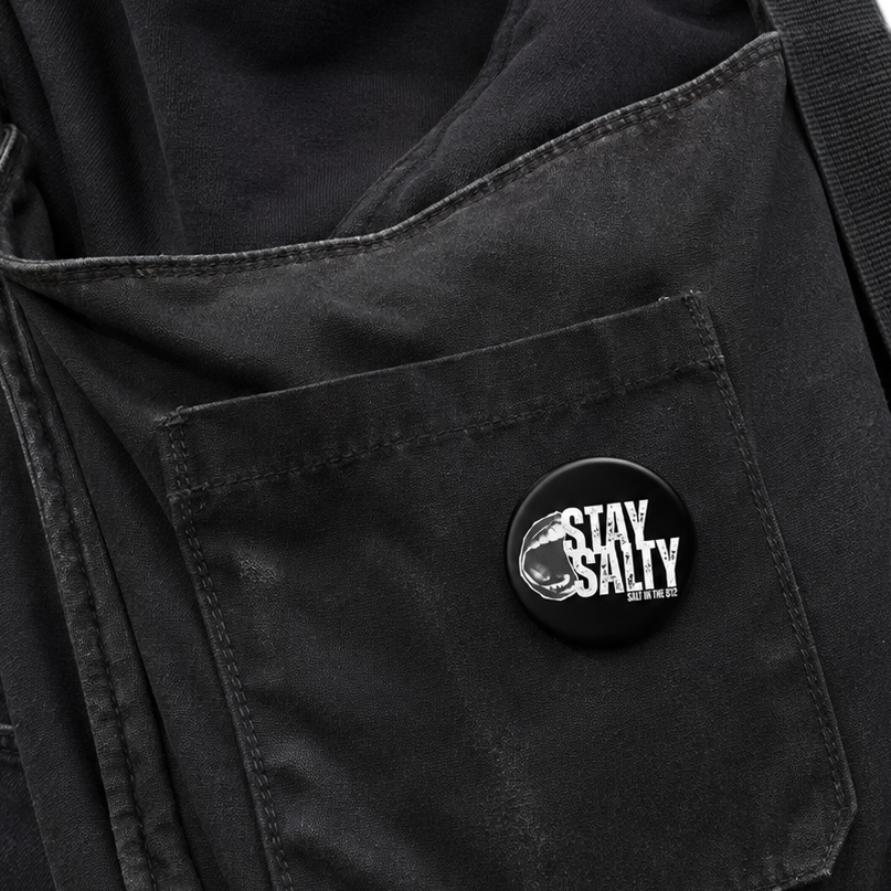 A close-up of a black canvas shoulder bag worn over a black shirt, featuring a front pocket with a circular black button that reads 'STAY SALTY' in white text.