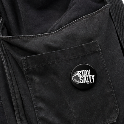 A close-up of a black canvas shoulder bag worn over a black shirt, featuring a front pocket with a circular black button that reads 'STAY SALTY' in white text.