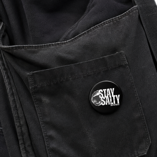 A close-up of a black canvas shoulder bag worn over a black shirt, featuring a front pocket with a circular black button that reads 'STAY SALTY' in white text.